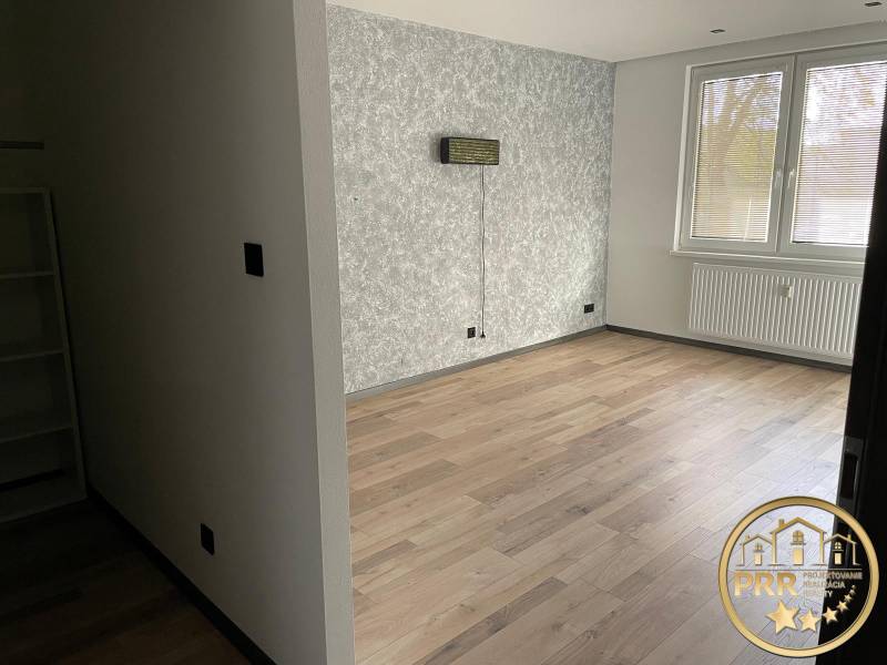Prázdná místnost v 3-pokojovém bytě s dřevěnou podlahou a strukturální stěnou.