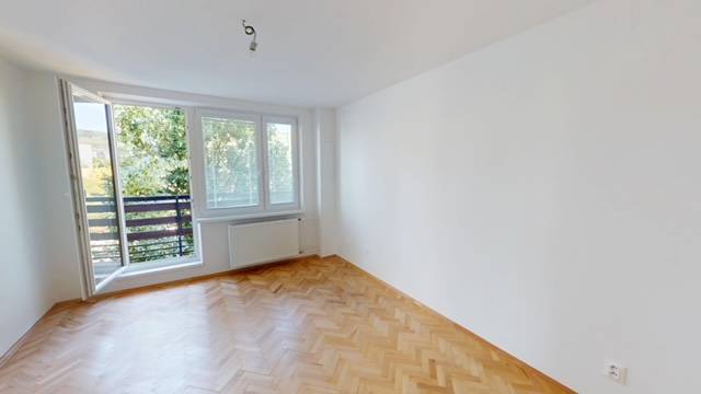 Prostorný pokoj v 2pokojovém bytě s dřevěnou podlahou a velkým oknem na balkon.