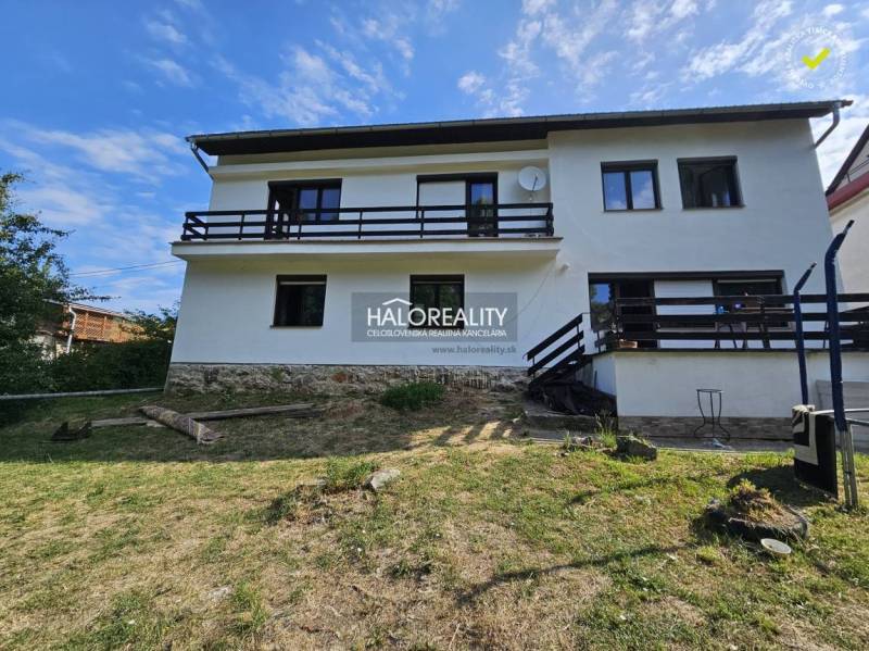 Rodinný dům v Banské Štiavnici s dřevěným balkonem na svahu a travnatým pozemkem.