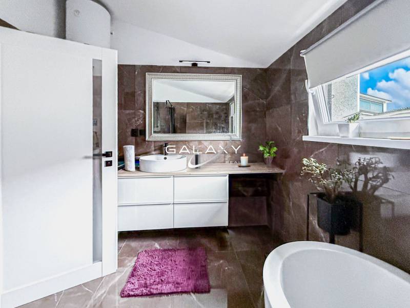 Koupelna v rodinném domě s vanou, umyvadlem a oknem, dekorativní rostliny.