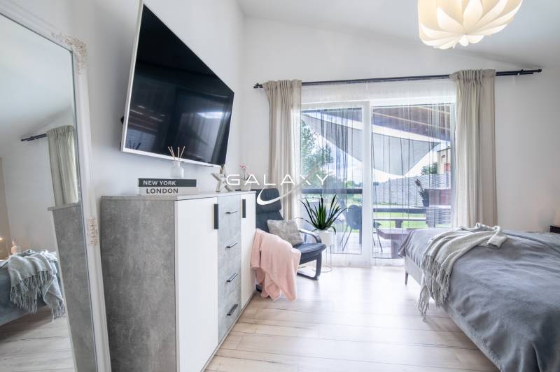 Světlá ložnice v rodinném domě s televizí, zrcadlem a balkonem.