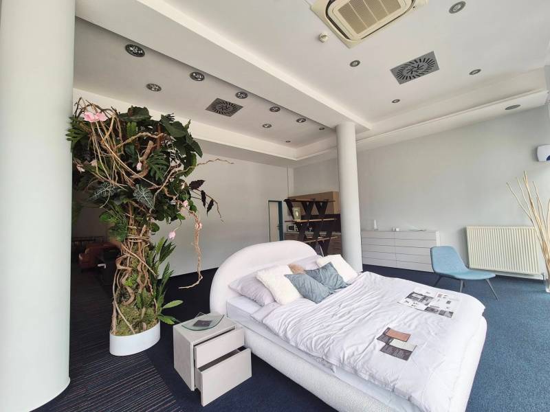 Postel s polštáři a rostlinami v obchodních prostorách, vedle noční stolek a křeslo.