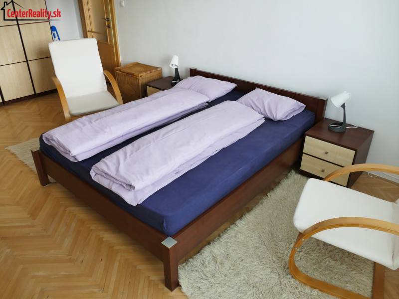 Ložnice ve 3pokojovém bytě s dřevěnou postelí, židlí a bílým kobercem.