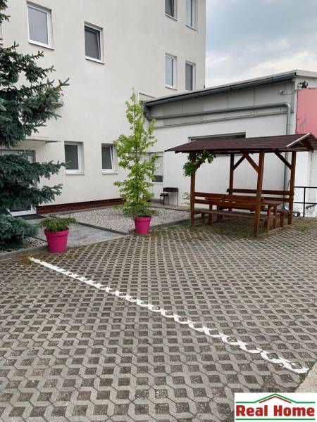 Dvorec s dlažbou, dřevěné sezení a rostliny v růžových květináčích na ulici Šamorínska.
