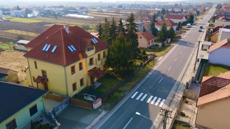Budova na ulici Chrabrany v Topoľčanoch, obklopená domy a stromy, u hlavní silnice.