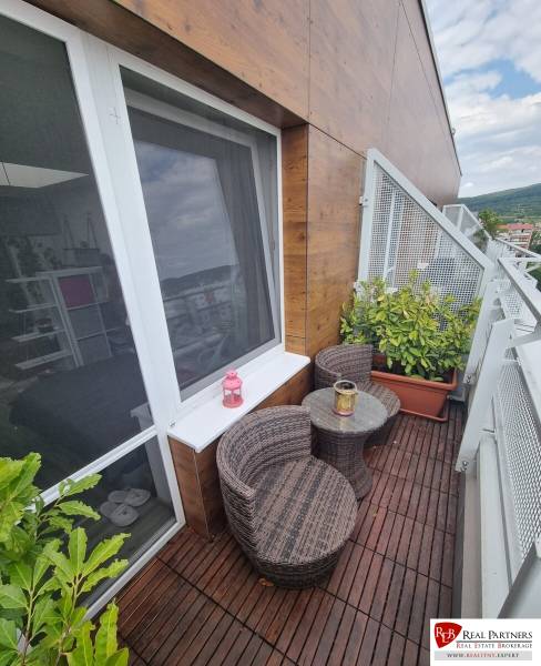 Balkon 1-pokojového bytu s ratanovým nábytkem a rostlinami u posuvného okna.