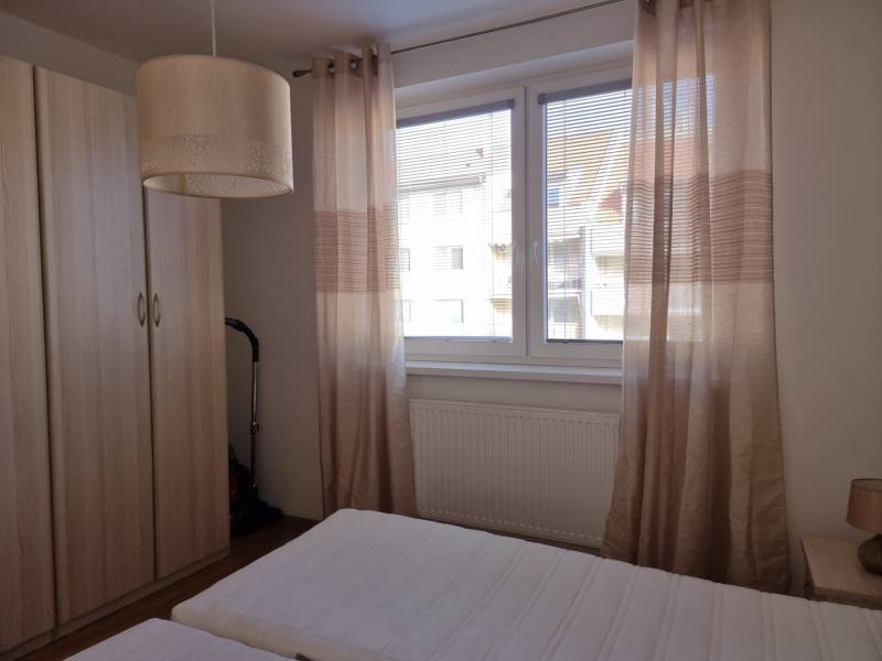 Ložnice v dvoupokojovém bytě s postelí, skříní a oknem s béžovými závěsy.