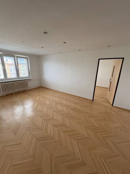 Pokoj s parketovou podlahou, bílými stěnami a okny, součást dvoupokojového bytu.