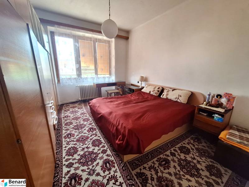Ložnice v dvoupokojovém bytě s dvojlůžkem, koberec, skříně, okno s radiátorem.
