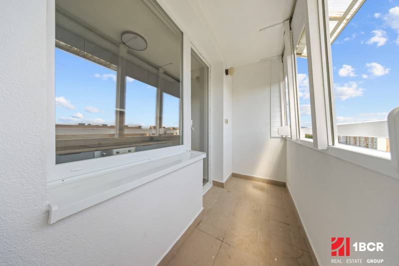 Světlý balkon 2-pokojového bytu s výhledem na okolní střechy a modrou oblohu.