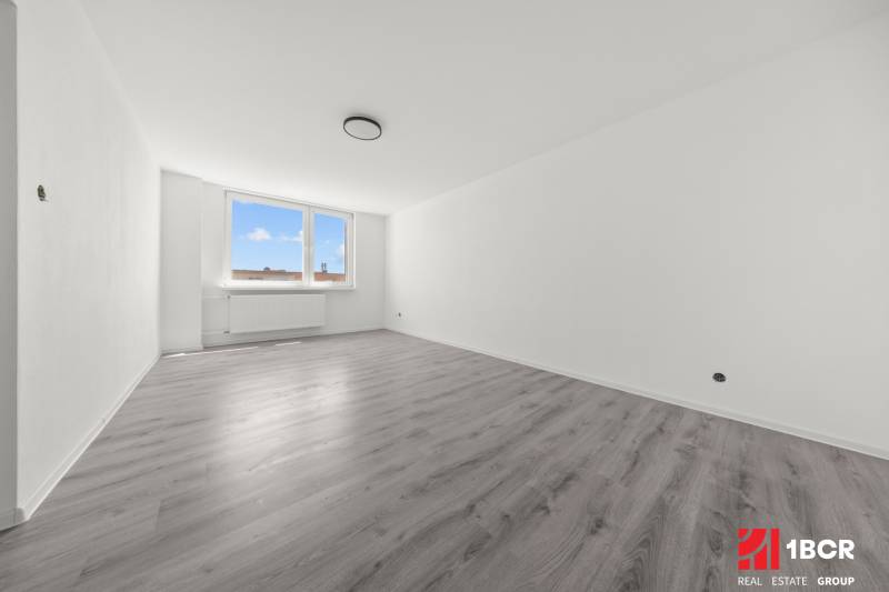 Prostorný pokoj s dřevěnou podlahou a velkým oknem v 2pokojovém bytě.