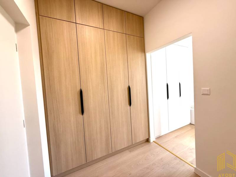 Garsonka s dřevěnými vestavěnými skříněmi a bílými stěnami působí útulně.