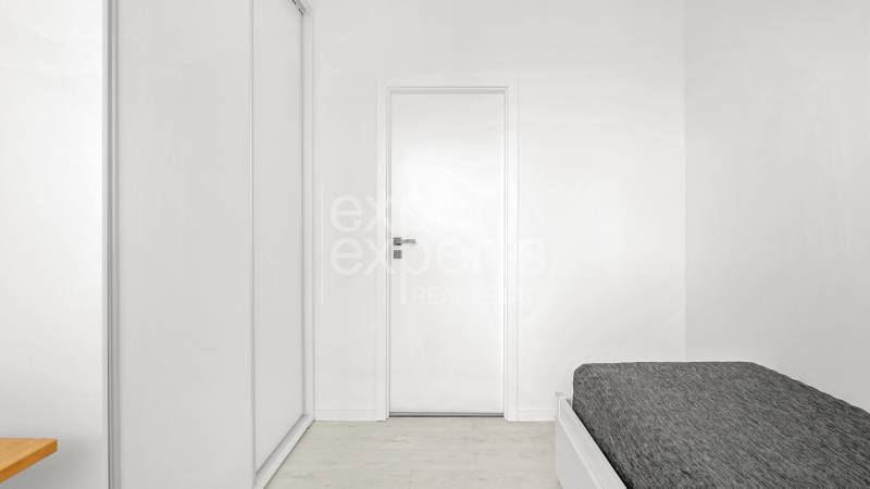 Světlá ložnice s bílou šatní skříní, dřevěným stolkem a postelí v třípokojovém bytě.
