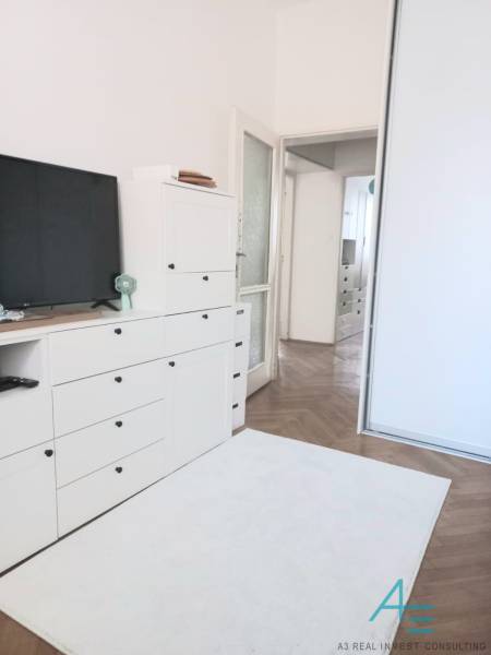 Bílý obývací pokoj ve 4pokojovém bytě s TV, skříňkami a dřevěnou podlahou.