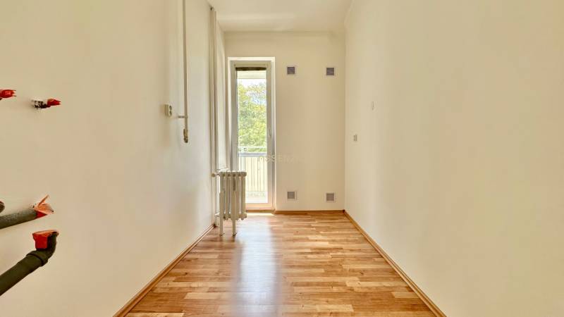 Pokoj s dřevěnou podlahou a balkónem ve 3-pokojovém bytě, bílá stěna, radiátor.