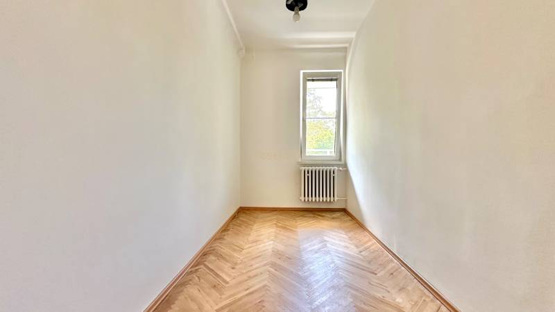 Pokoj se světlými stěnami, dřevěnou podlahou, radiátorem a oknem ve 3-pokojovém bytě.