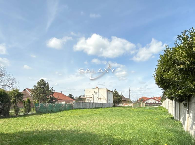 Pozemek k bydlení ve Sládkovičově, tráva, ploty, stromy, budovy v pozadí, oblačná obloha.