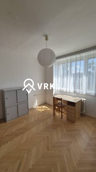 Osvětlený pokoj v 2-pokojovém bytě se stolem, židlí a skříňkou na parketové podlaze.