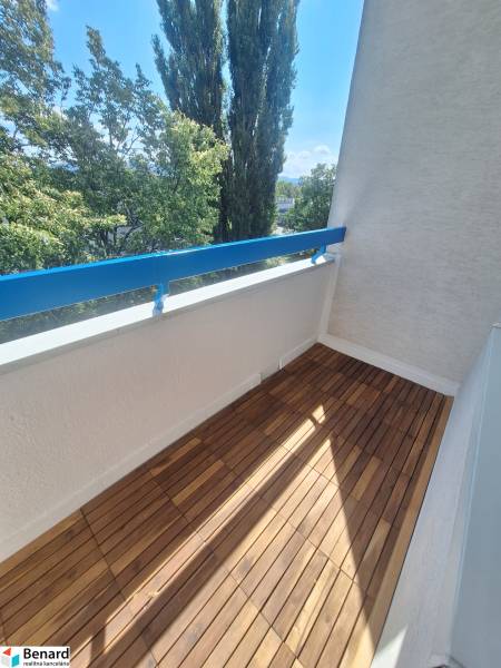 Balkon s dřevěnou podlahou a výhledem na stromy v 2-pokojovém bytě.