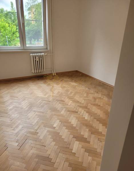 Pokoj v 3-pokojovém bytě s dřevěnými parketami, velké okno, radiátor pod ním.