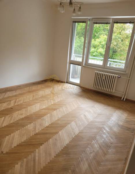 Pokoj ve 3-pokojovém bytě s dřevěnou podlahou a balkonovými dveřmi.