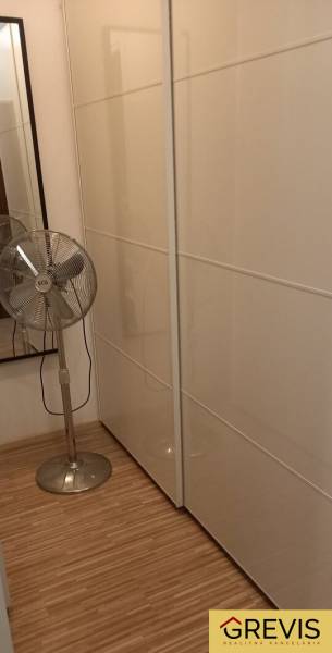 Šatník v 2-pokojovém bytě s bílými posuvnými skříněmi a stojanovým ventilátorem.