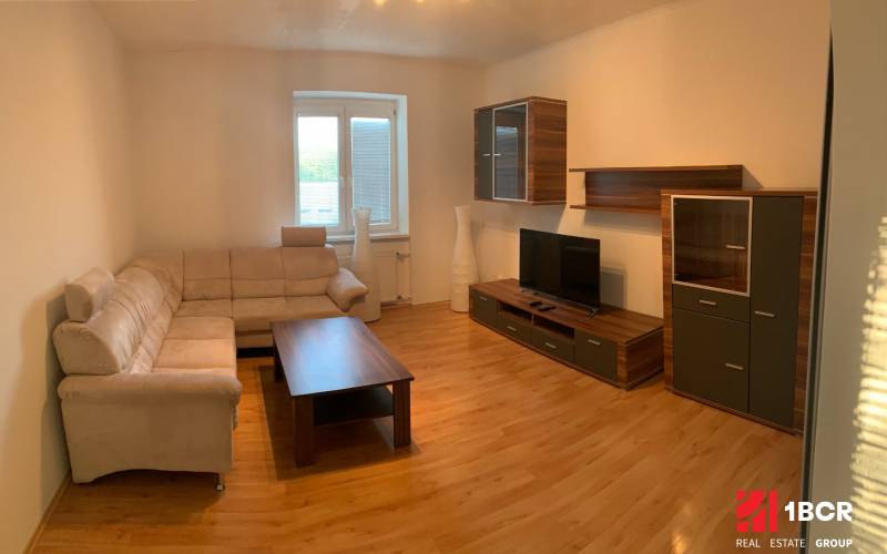 Obývací pokoj s béžovou sedačkou, TV a dřevěným dekorem v 2pokojovém bytě.