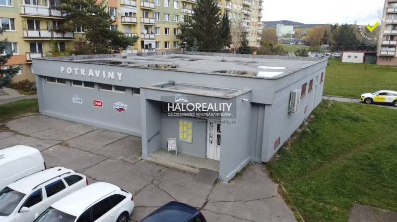 Budova obchodního prostoru v Rimavské Sobotě, obklopená paneláky a zaparkovanými auty.