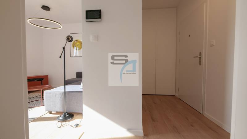 Interiér 2-pokojového bytu s podlahou s dřevěným dekorem, kruhovým lustrem a šedou pohovkou.