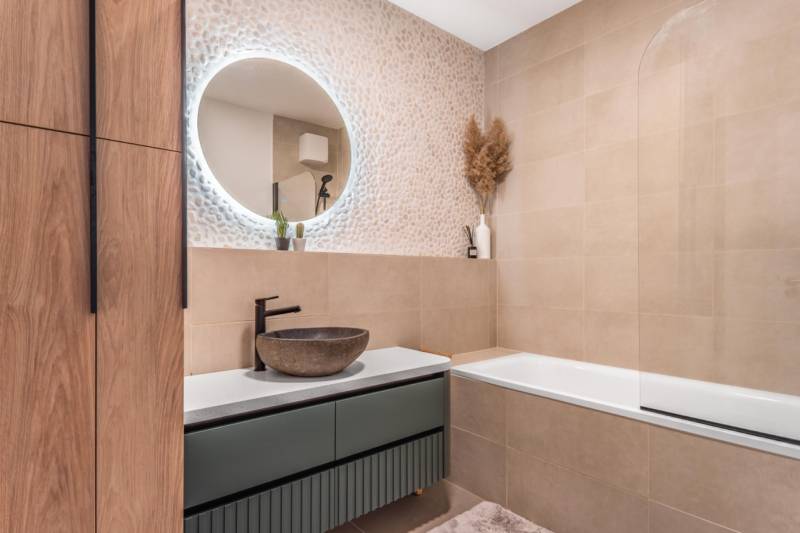 Koupelna v 3-pokojovém bytě se vzorovanou stěnou, kulatým zrcadlem a vanou.