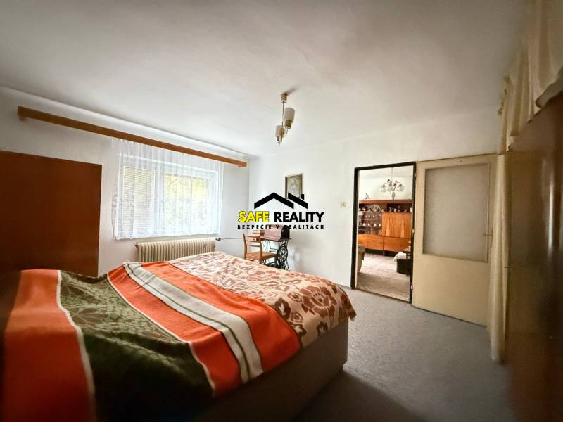 Ložnice v rodinném domě se světlým kobercem, oknem a postelí s dekou.