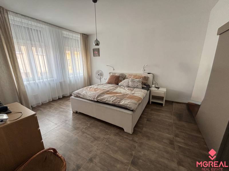 Ložnice v dvoupokojovém bytě s velkými okny, postelí a podlahou s dřevěným dekorem.