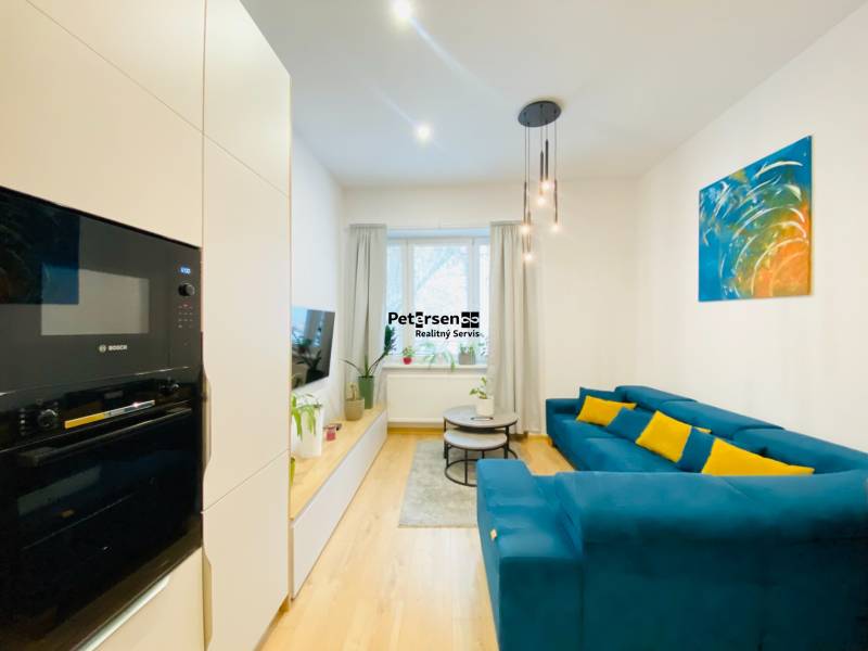 Obývací pokoj s modrou pohovkou, barevným obrazem a podlahou s dřevěným dekorem v 2pokojovém bytě.