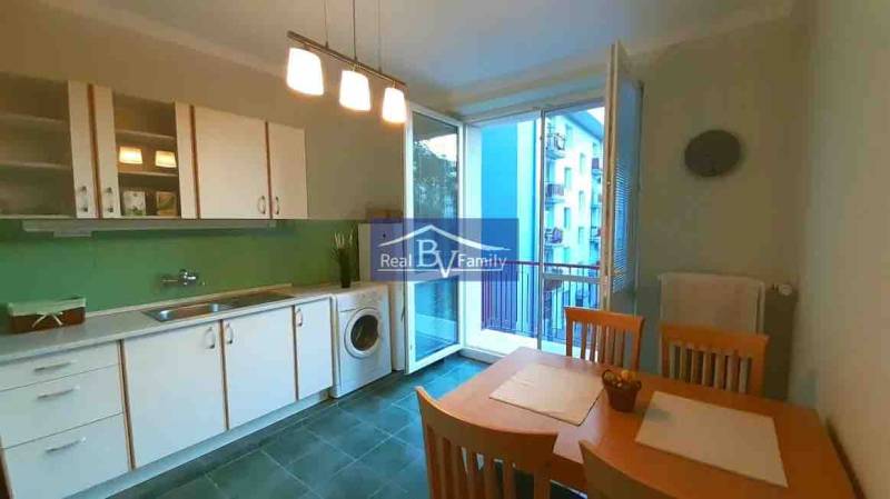 Kuchyně v 2pokojovém bytě s bílými skříňkami, jídelním stolem a balkónem.