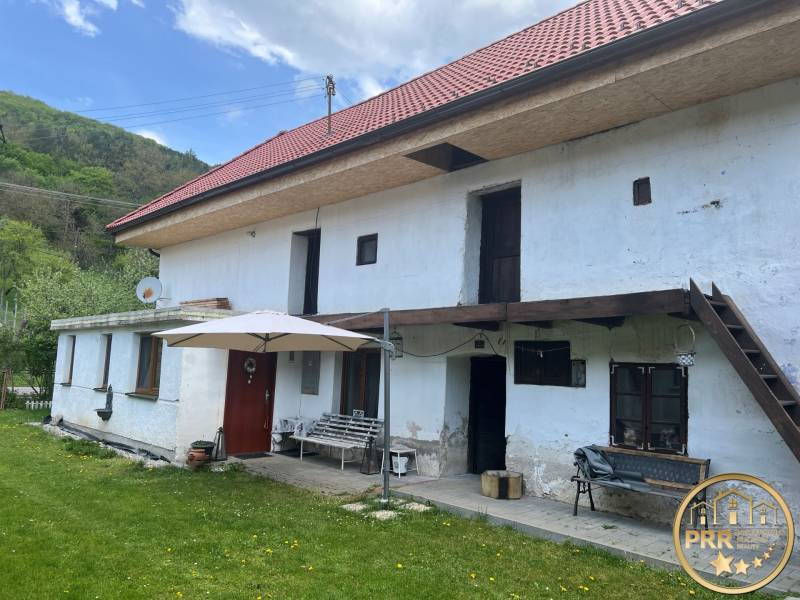 Venkovská chata s červenou střechou a terasou, obklopená zeleným trávníkem a přírodou.