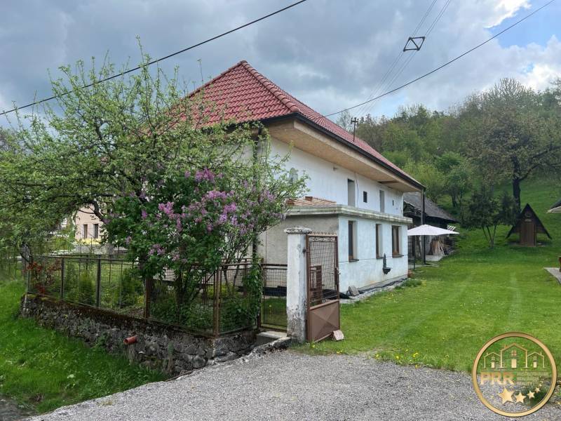 Venkovská chata obklopená zelení, s rustikální střechou, kvetoucí zahradou a dřevěným altánkem.