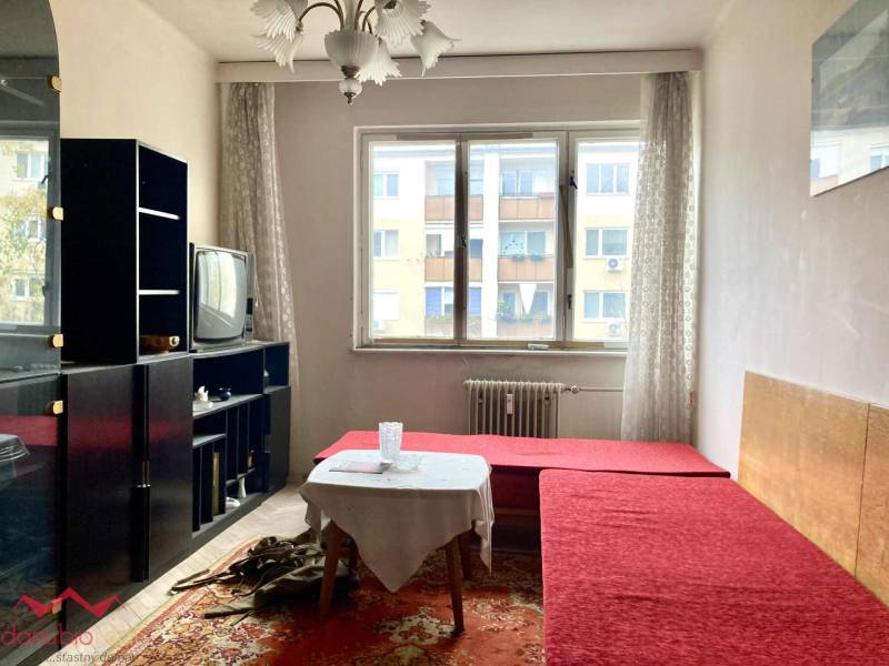 Obývací pokoj ve 3-pokojovém bytě s červenou pohovkou, koberec, starší televizor, prosvětlený prostor.