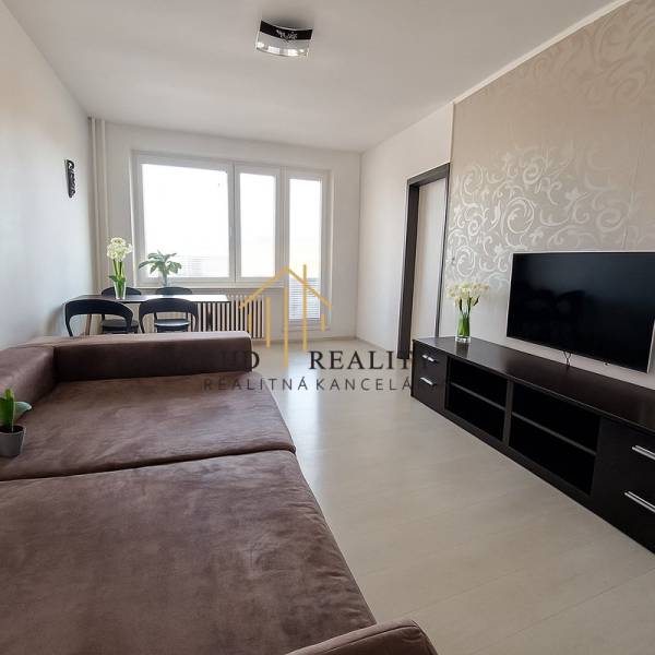 Obývací pokoj v 3-pokojovém bytě s velkou sedačkou, TV a podlahou s dřevěným dekorem.