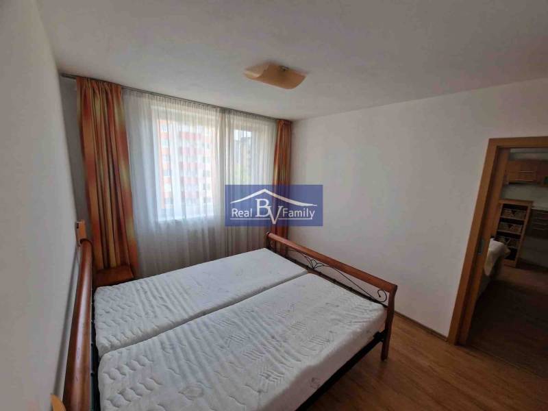 Ložnice v dvoupokojovém bytě s postelí, závěsy a podlahou s dřevěným dekorem.