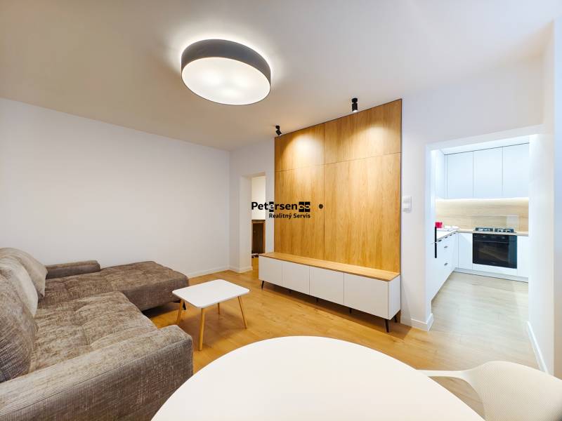 Obývací pokoj v 2-pokojovém bytě s podlahou s dřevěným dekorem, vstupem do bílé kuchyně.