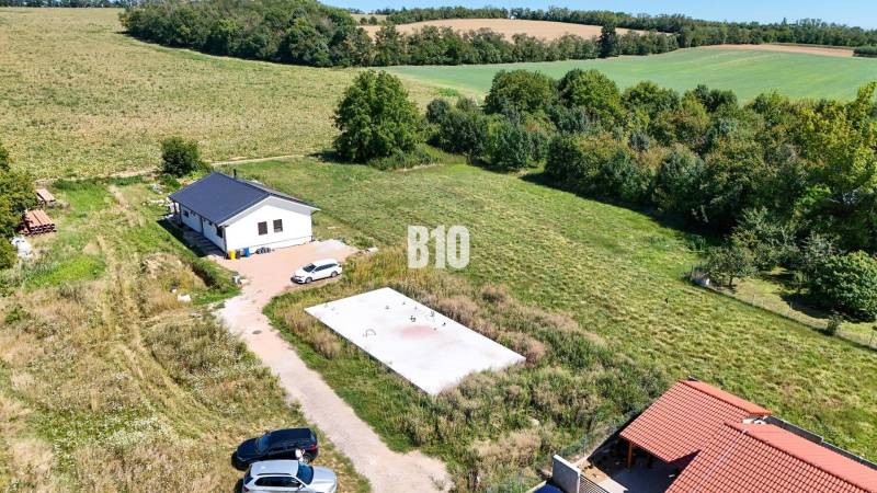 Záběr na pozemek pro bydlení v Čeľadicích zahrnuje dům, zeleň a auta.