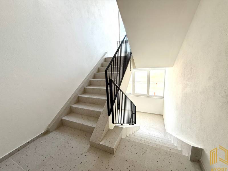 Schodiště v bytovém domě s oknem, bílé stěny a dlažba.