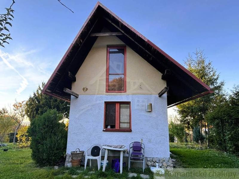 Chata v Sološnici se strmou střechou, okna, příroda a plastové židle.