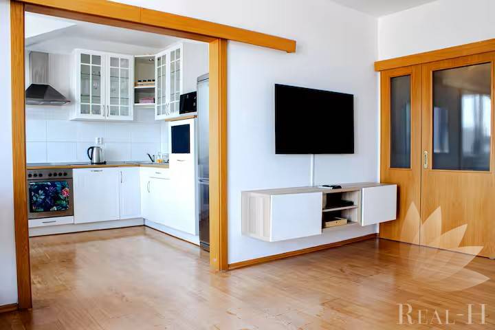 Obývací pokoj v 3-pokojovém bytě s průchodem do kuchyně a nástěnnou televizí.