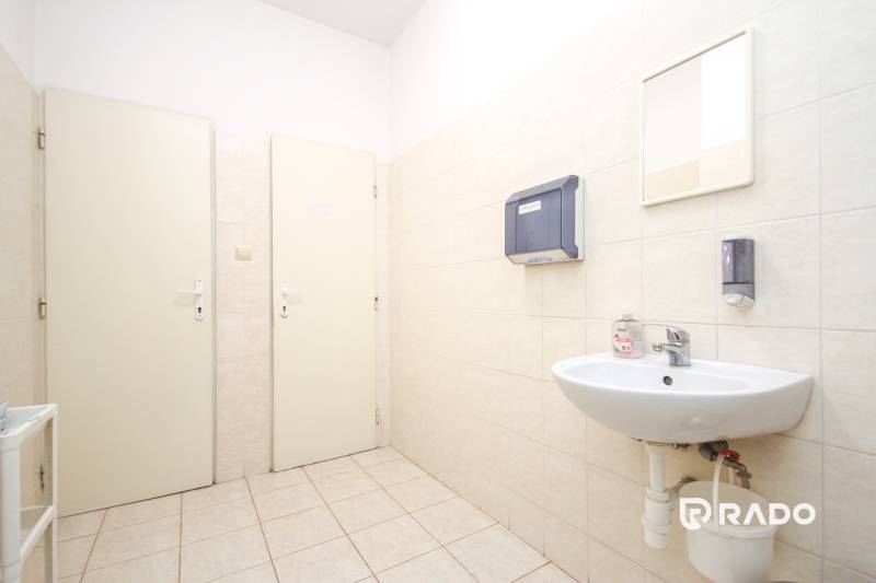 Kancelář s umyvadlem, dávkovači a zrcadlem. Obklady bílé, podlaha světlá.