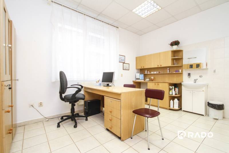 Kancelář s počítačem, stolem a židlí, světlý interiér, police a umyvadlo.