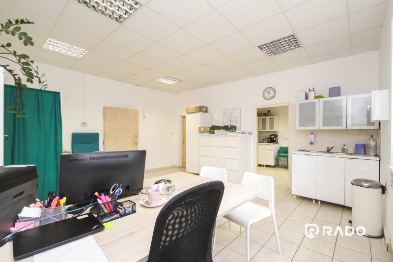 Kancelář s nábytkem, počítačem a kuchyňkou, na podlaze jsou dlaždice.