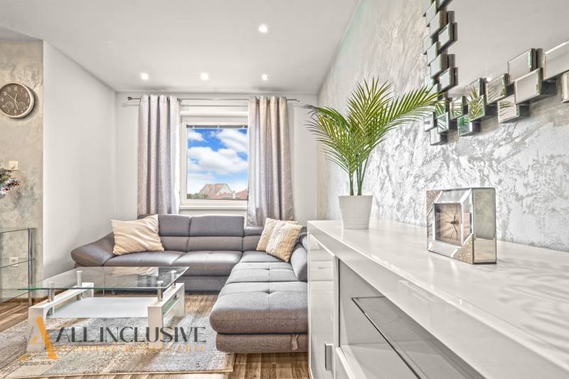 Obývací pokoj s šedou sedačkou, skleněným stolkem a podlahou s dřevěným dekorem.