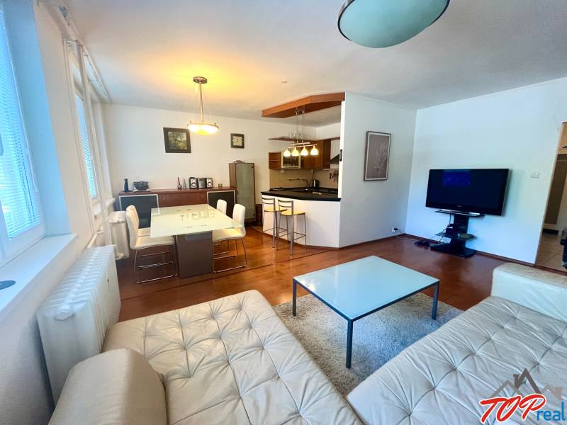 Obývací pokoj s kuchyní v 3pokojovém bytě, vybavení zahrnuje sedačku, stůl a televizi.