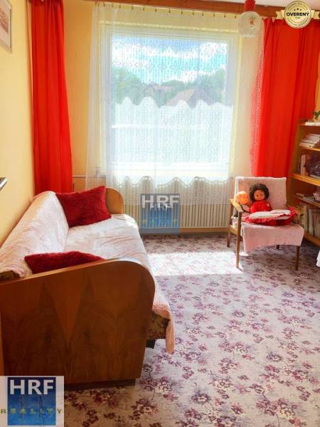 Pokoj v rodinném domě s květovaným kobercem, pohovkou a poličkami.